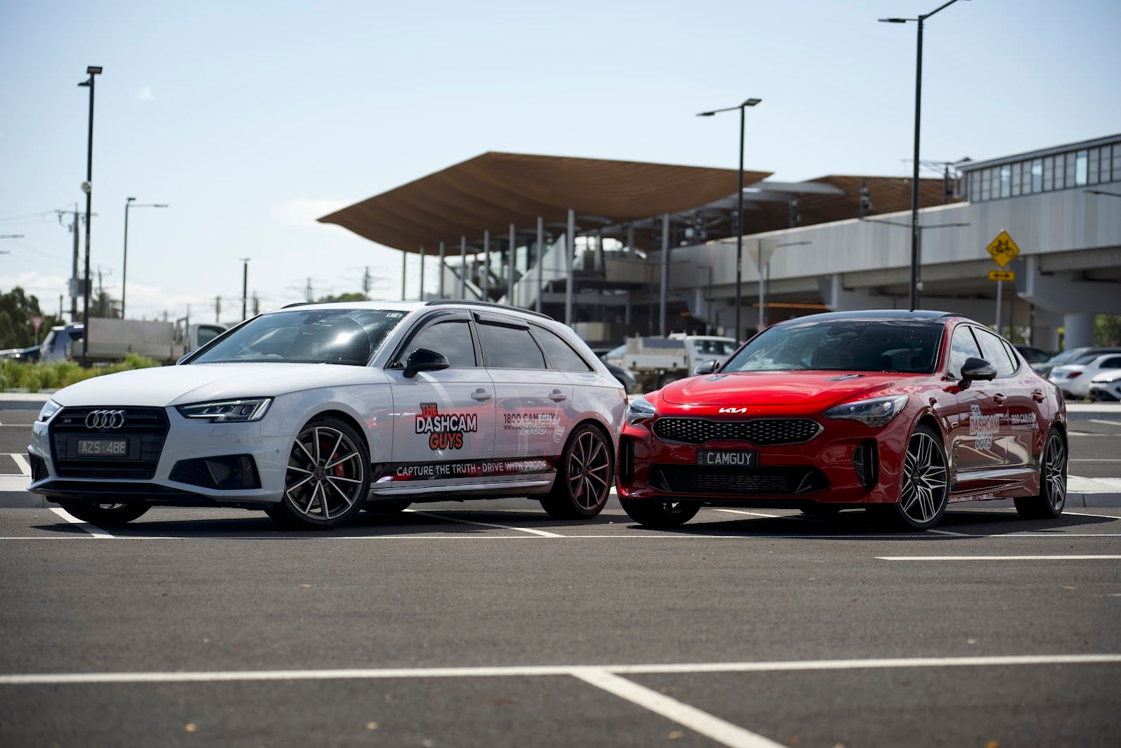 1 Audi and 1 Kia cars, one white and one red, parked on a tarmac surface with a building in the background.